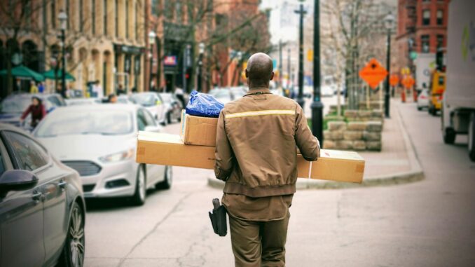 Zusteller Ein Paketbote in brauner Uniform trägt mehrere Pakete auf einer belebten Stadtstraße.